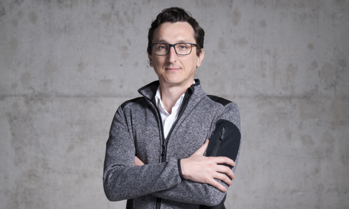 Ein Mann steht vor einer grauen Wand, trägt eine graue Jacke mit schwarzen Details über einem schwarzen Hemd und hat die Arme verschränkt. Er trägt eine Brille und schaut freundlich in die Kamera.