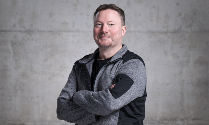 Ein Mann steht vor einer grauen Wand, trägt eine graue Jacke mit schwarzen Details über einem schwarzem T-Shirt und hat die Arme verschränkt. 