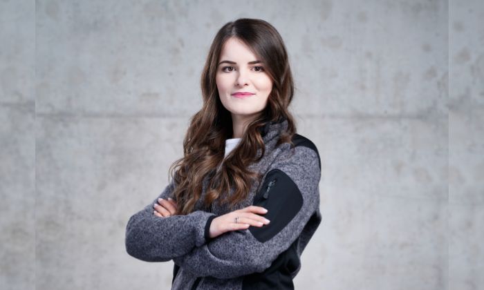 Eine Frau steht vor einer grauen Wand, trägt eine graue Jacke mit schwarzen Details über einem weißen T-Shirt und hat die Arme verschränkt. 